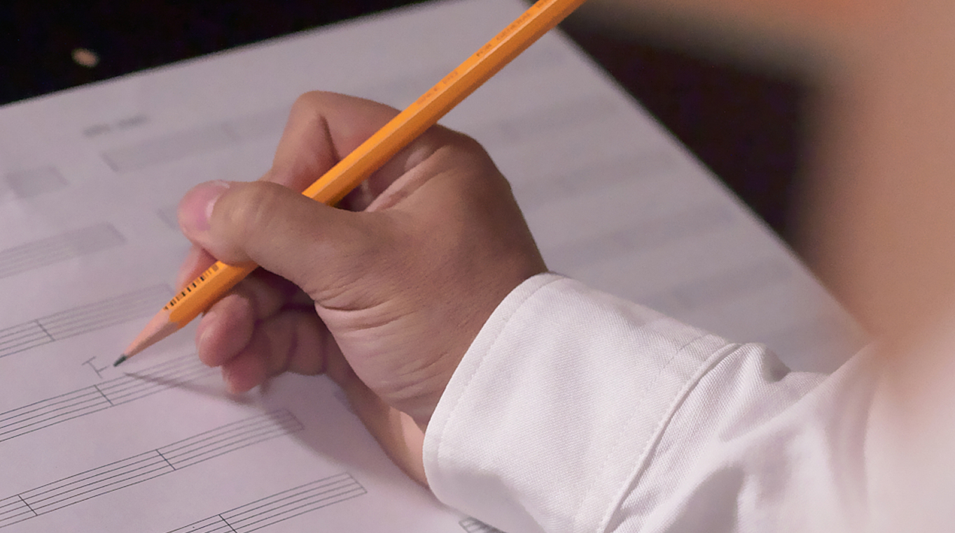 A close-up of a hand holding an orange pencil, writing on a sheet of music notation.