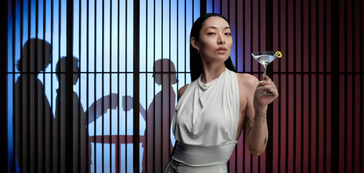 Rina Sawayama holds a martini in front of a jail cell, blending elegance with a hint of rebellion.