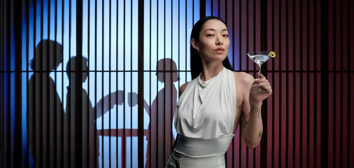 Rina Sawayama holds a martini in front of a jail cell, blending elegance with a hint of rebellion.