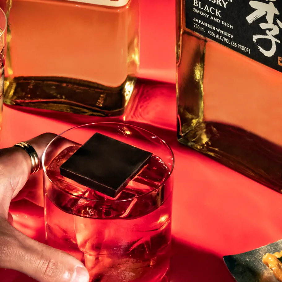 A hand reaches for a glass of whiskey with a dark ice cube, surrounded by other drinks and Toki black bottle on a red background.