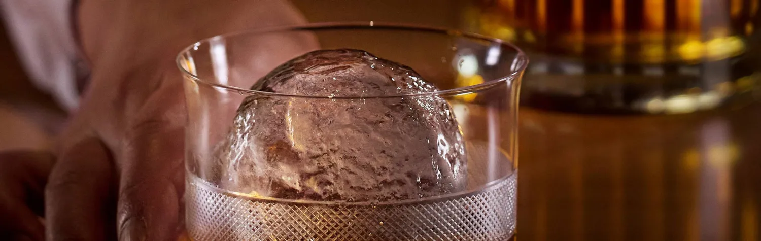 A hand placing a glass of whiskey with a large ice sphere on a wooden surface, with a blurred bottle in the background.