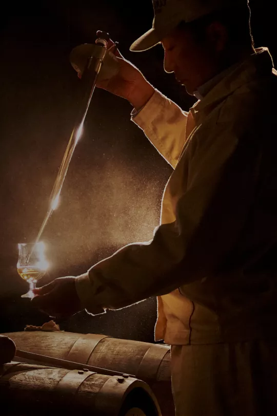 Image a of a man decanting whisky into a glass standing over whisky barrels
