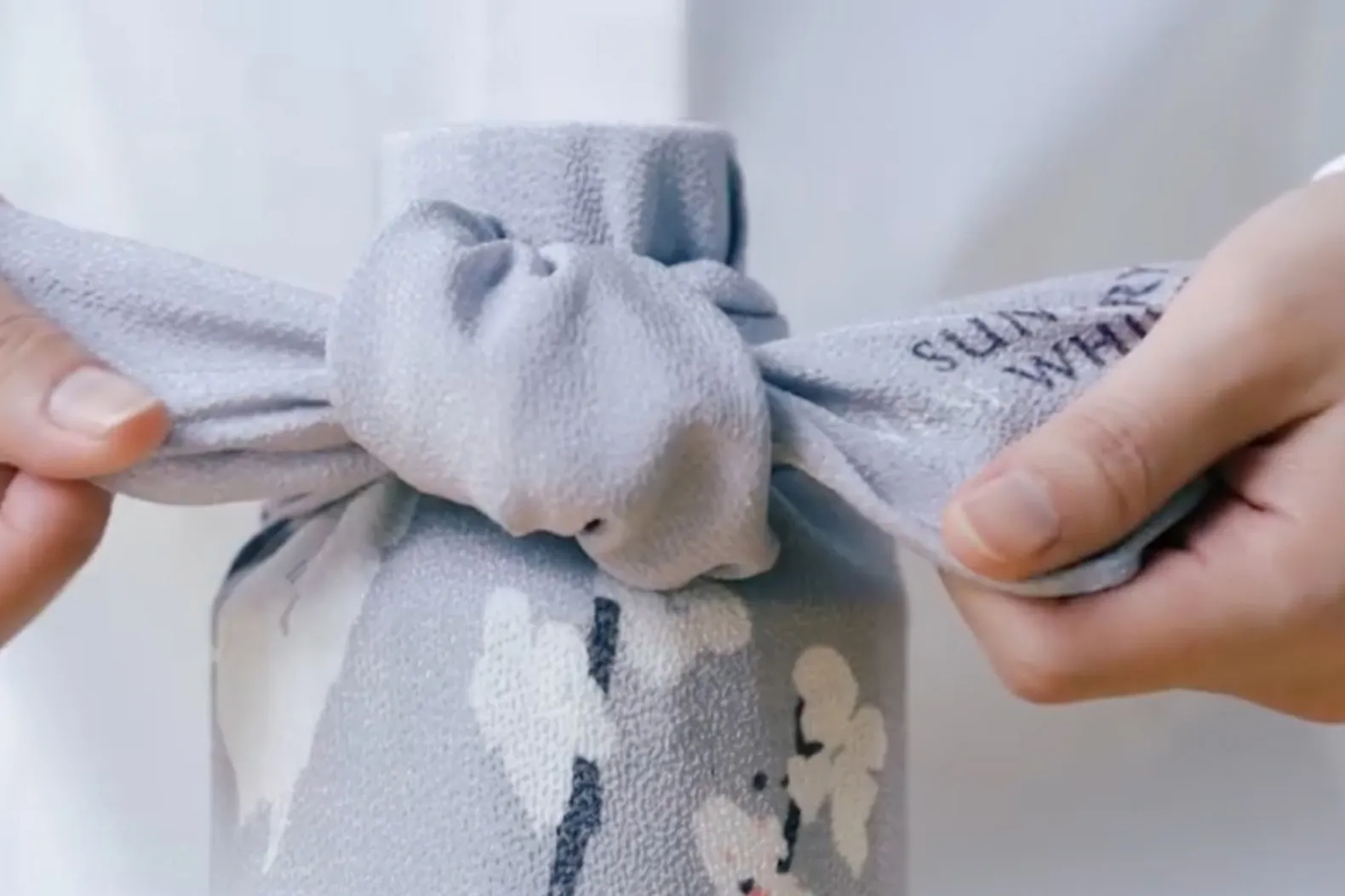 Hands tying a knot with a gray fabric featuring partial text and design.