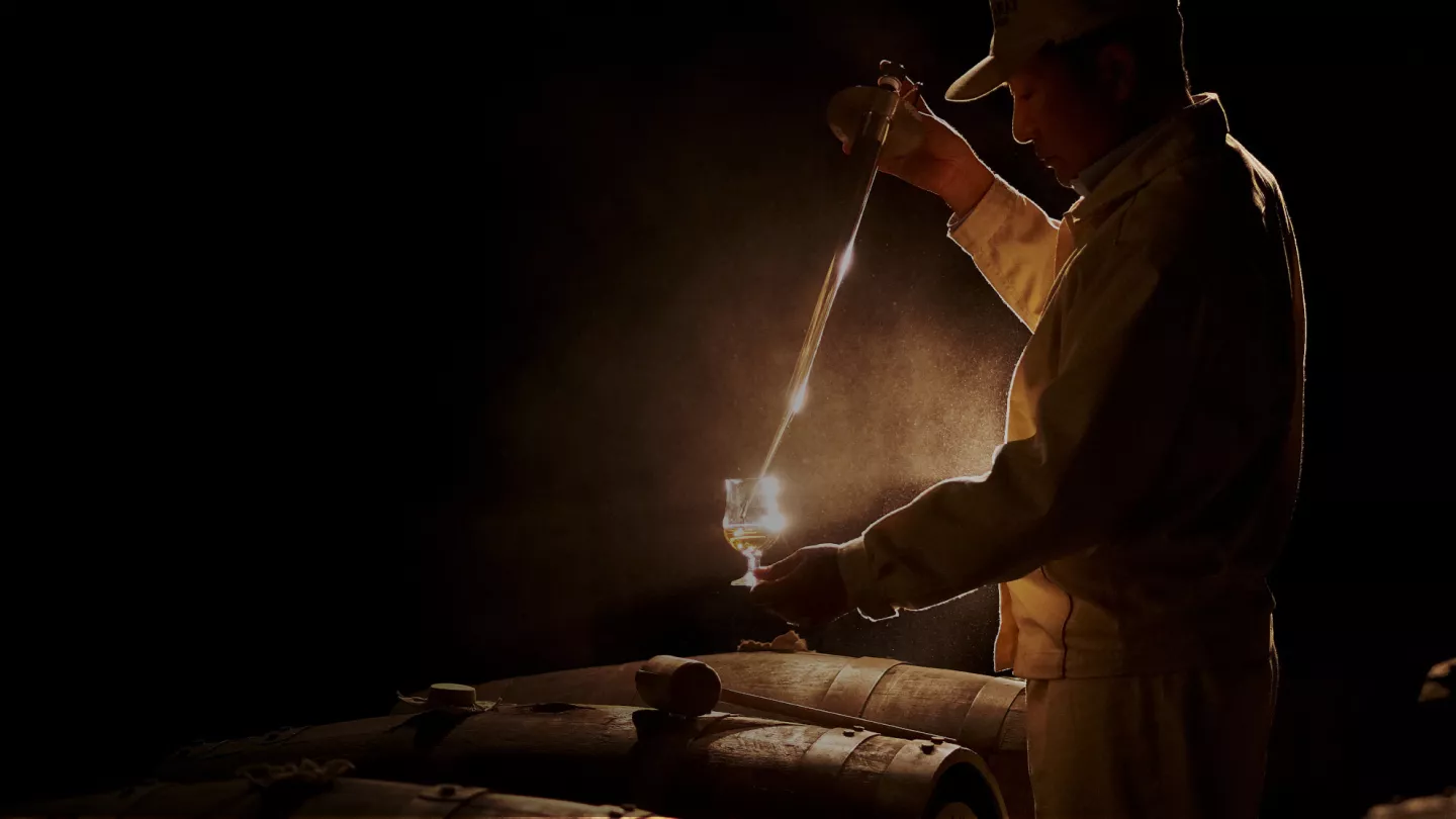 Image a of a man decanting whisky into a glass standing over whisky barrels