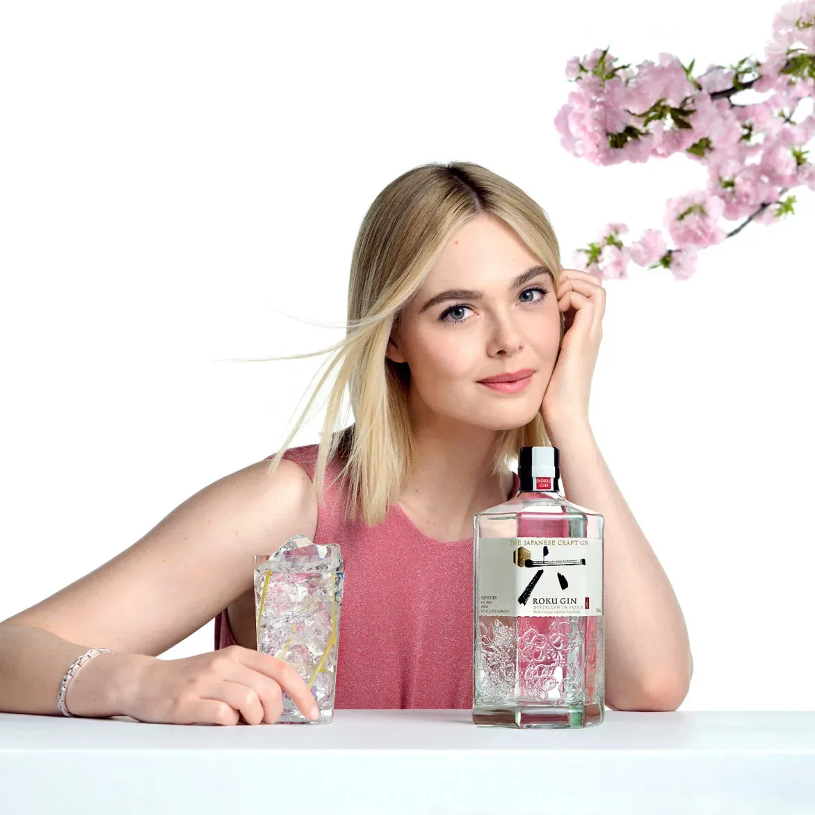 A woman elegantly holds a bottle of Roku Gin whiskey while standing beside a delicate cherry blossom.