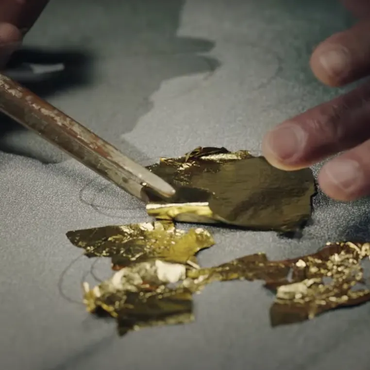 A person skillfully cuts gold foil with a knife, showcasing precision and attention to detail in the crafting process.