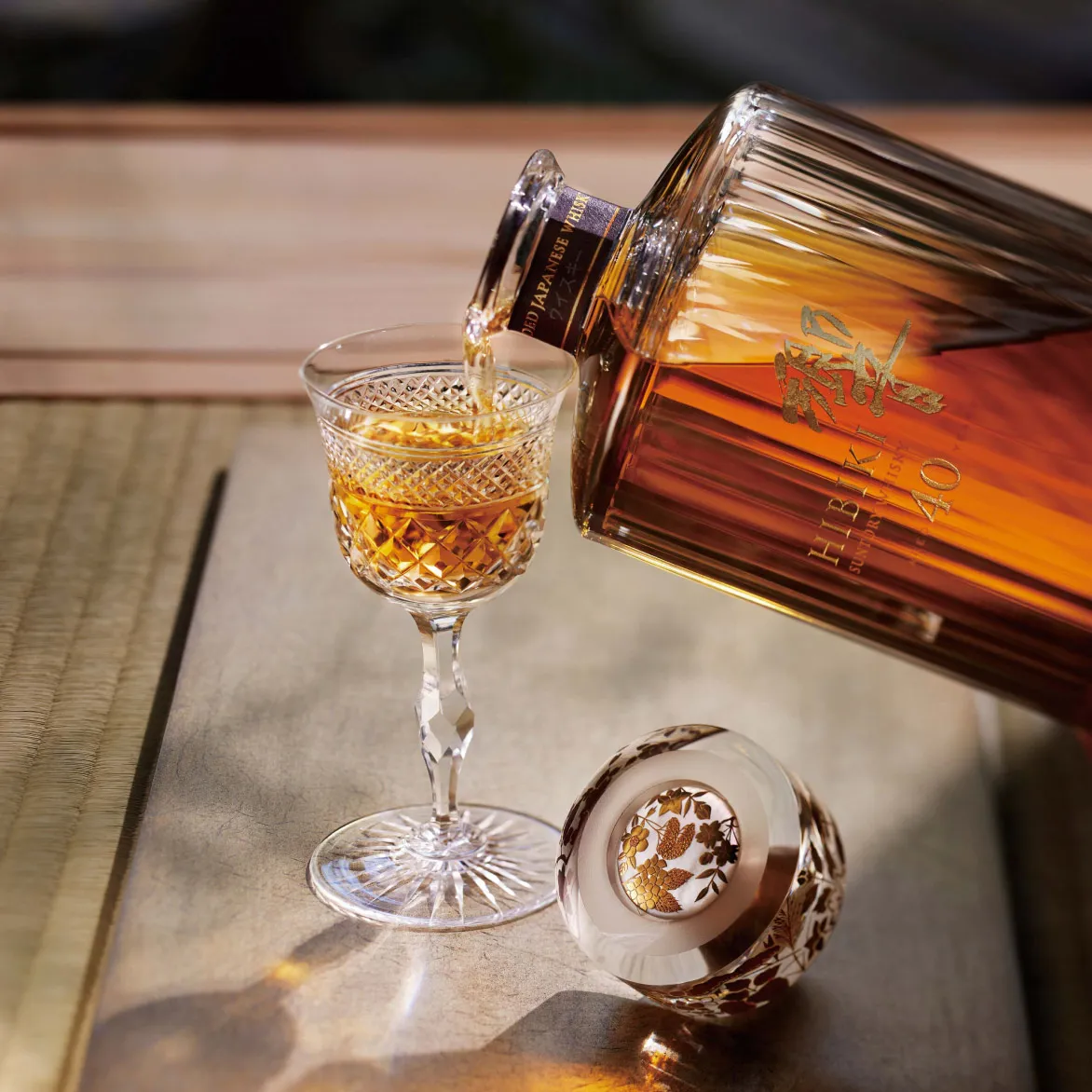 Amber liquor being poured into a crystal glass on a wooden table with a capped bottle beside it.