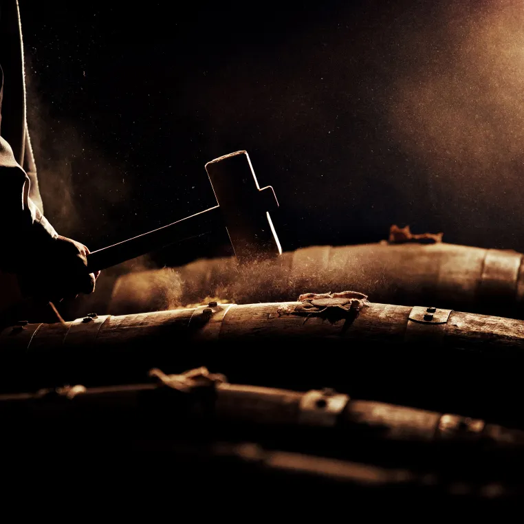 A person hammering the top of stacked wooden barrels in dim lighting. Dust is visible around the hammer.
