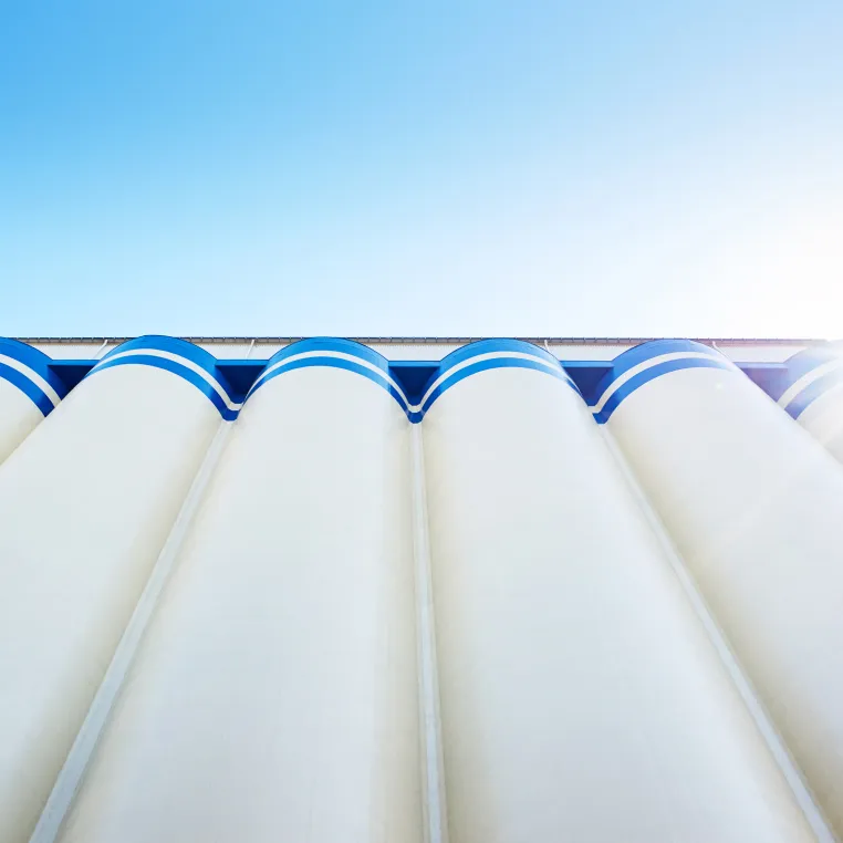 Looking up at tall white silos with blue trim against a bright blue sky with sunlight peeking through.