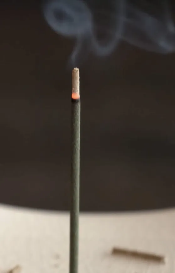 A lit incense stick with a glowing tip and delicate smoke rising against a blurred background.