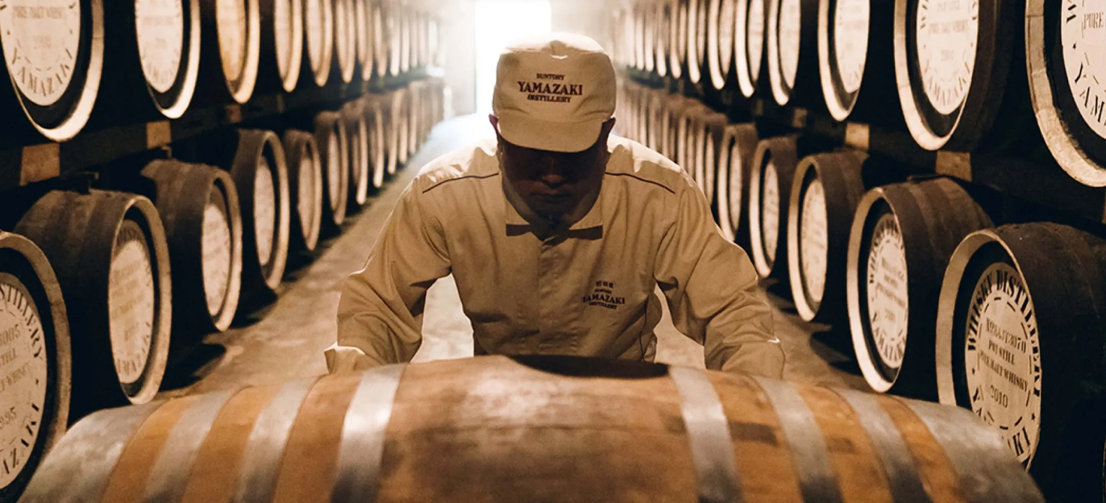 Worker in a distillery with rows of wooden barrels aging spirits.