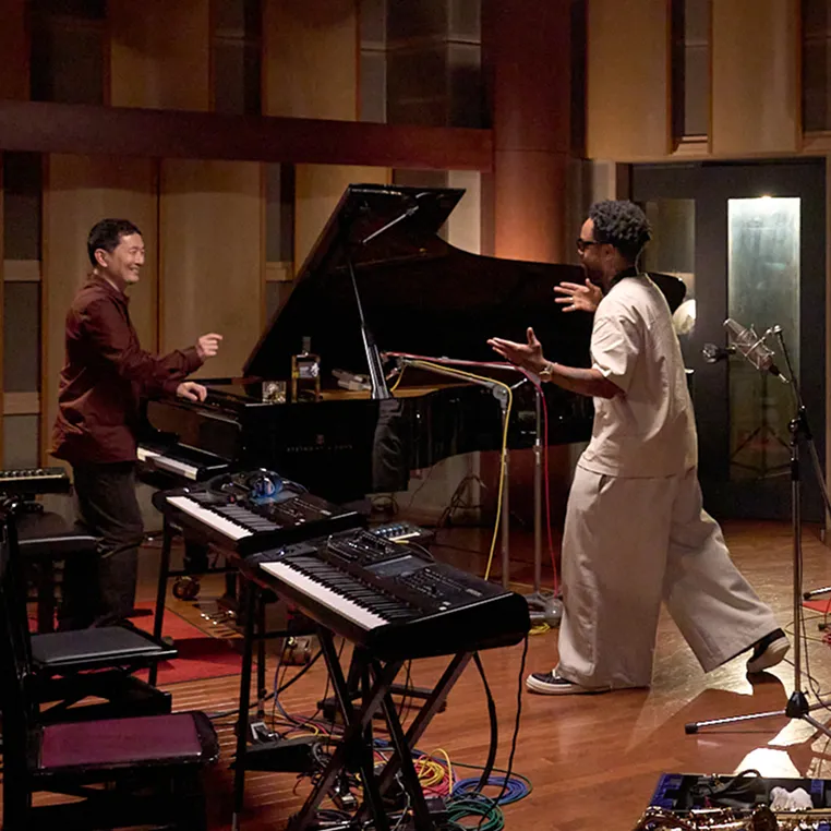 Two musicians engage passionately in a recording studio, surrounded by instruments, a grand piano, and colorful cables.