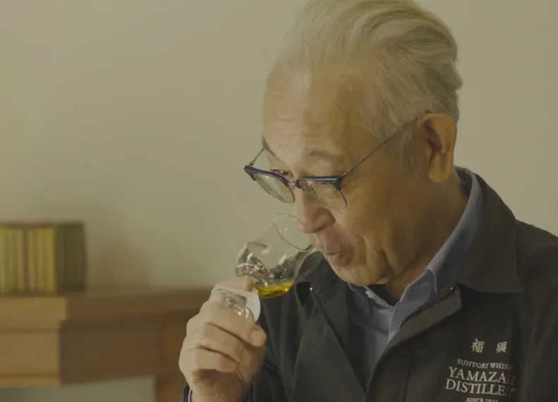 An elderly man holds a small glass of whiskey, observing its color thoughtfully, surrounded by a simple, elegant setting.