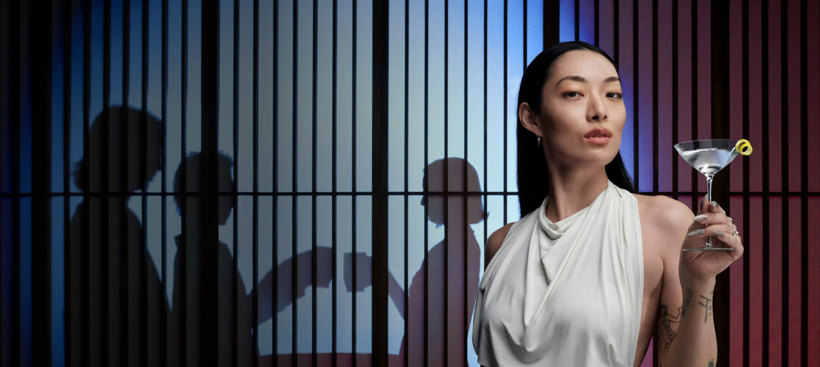 Rina Sawayama holds a martini in front of a jail cell, blending elegance with a hint of rebellion.